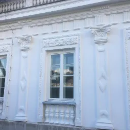 Biała, odnowiona elewacja budynku z dekoracyjnymi kolumnami, ornamentami nad oknami i balustradą pod oknami. Widoczne detale architektoniczne.