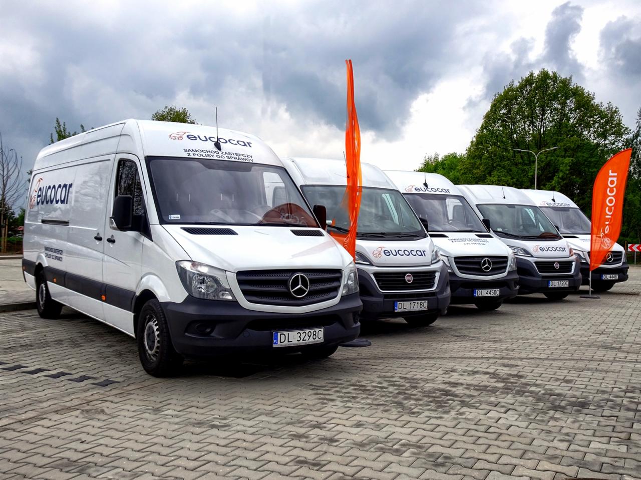 Rząd zaparkowanych, firmowych vanów z logo Euocar, stojących na brukowanym placu pod pochmurnym niebem, z pomarańczowymi flagami na skraju.