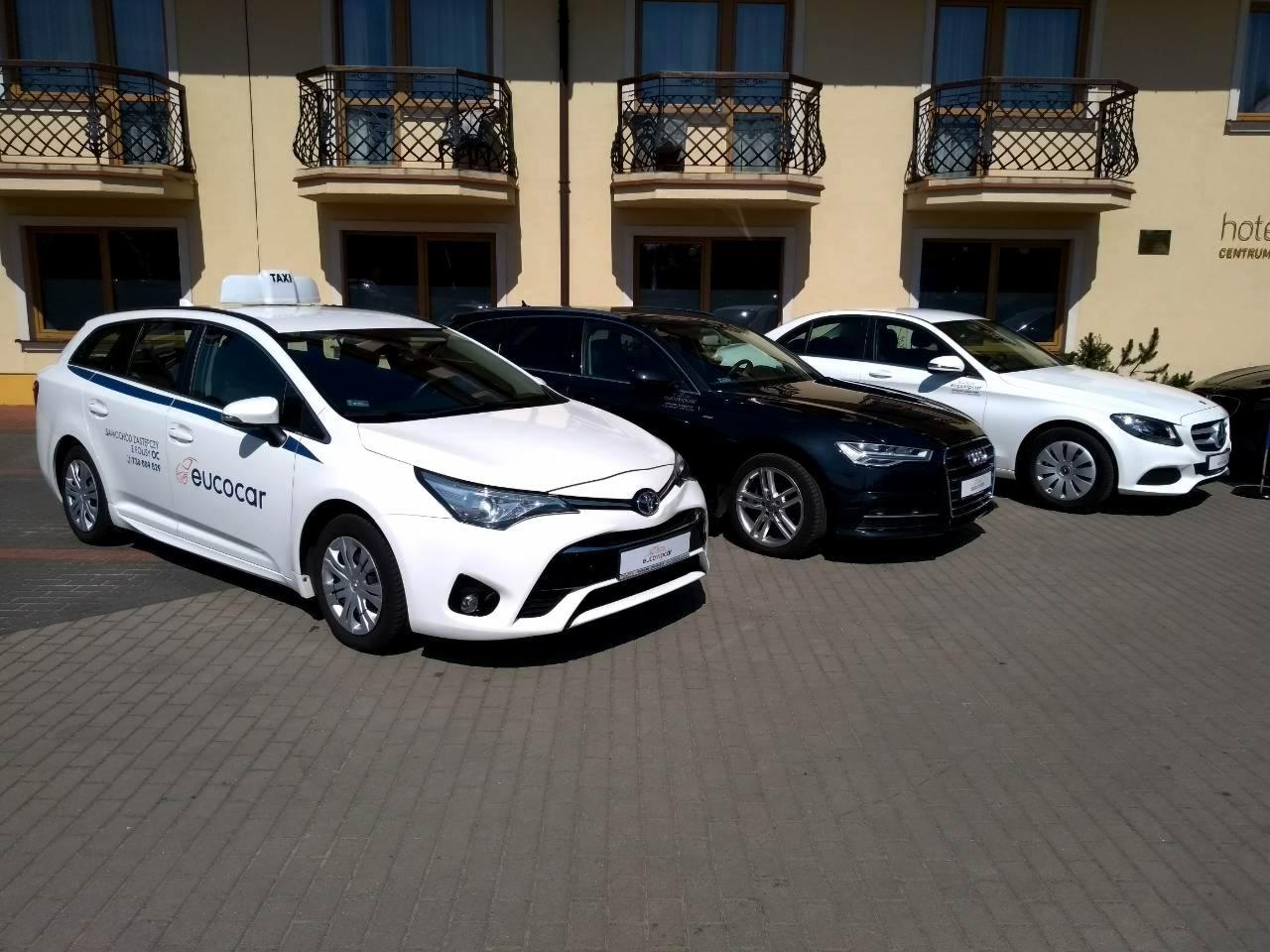 Flota samochodów osobowych różnych marek, w tym biała taksówka z napisem 'eucocar' na tle hotelu.