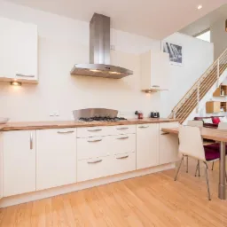 Jasna kuchnia w nowoczesnym dwupoziomowym apartamencie z drewnianymi podłogami, białymi szafkami i metalowym okapem nad kuchenką gazową, widok na schody i salon.