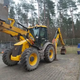 Żółta koparko-ładowarka JCB, lekko zabrudzona, stoi na kostce brukowej na tle drzew i metalowej siatki.