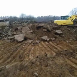 Żółta koparka na placu budowy, gruz i ziemia w tle z zalesionym terenem w pochmurny dzień.