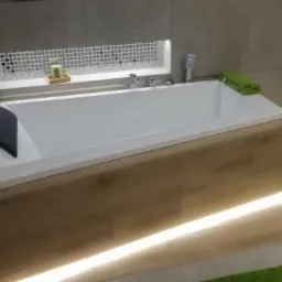 Nowoczesna, prostokątna wanna obudowana panelami imitującymi jasne drewno, z podświetleniem LED u dołu, na tle szarych płytek i mozaiki nad wanną, z zielonym ręcznikiem i akcesoriami.