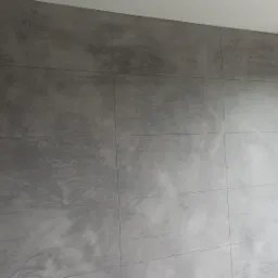 Ściana w Andrychowie wykończona szarą, dekoracyjną masą, imitującą beton architektoniczny, z widocznymi podziałami na płyty i oknem z roletą w tle.