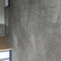 Ściana wykończona szarą, strukturalną okładziną imitującą beton, z widocznymi fugami, fragment szafki kuchennej z drewnianym blatem w lewym dolnym rogu.