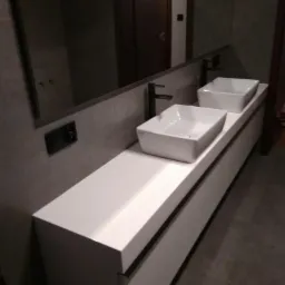 Nowoczesna łazienka z dwoma kwadratowymi umywalkami nablatowymi i czarnymi bateriami na długim, białym blacie z szafką podwieszaną, widok z niskiego kąta.