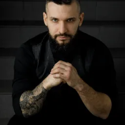 Portret brodatego mężczyzny z tatuażem na ramieniu, w ciemnej koszuli i kamizelce, siedzącego na schodach i patrzącego w obiektyw.