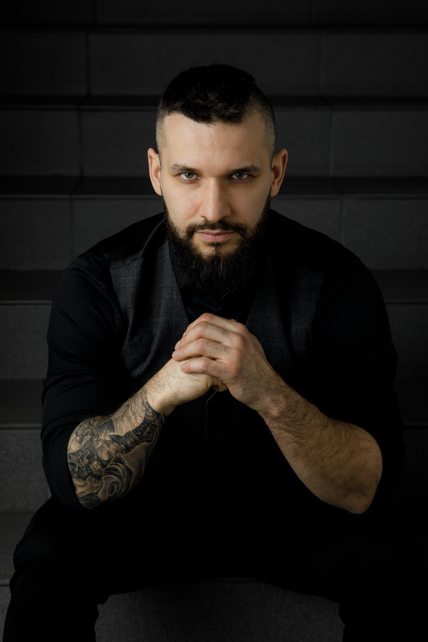 Portret brodatego mężczyzny z tatuażem na ramieniu, w ciemnej koszuli i kamizelce, siedzącego na schodach i patrzącego w obiektyw.