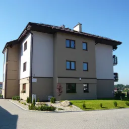 Trzypiętrowy budynek mieszkalny z beżowo-białą elewacją, brązowym dachem i oknami, otoczony zielenią, z samochodem zaparkowanym obok.