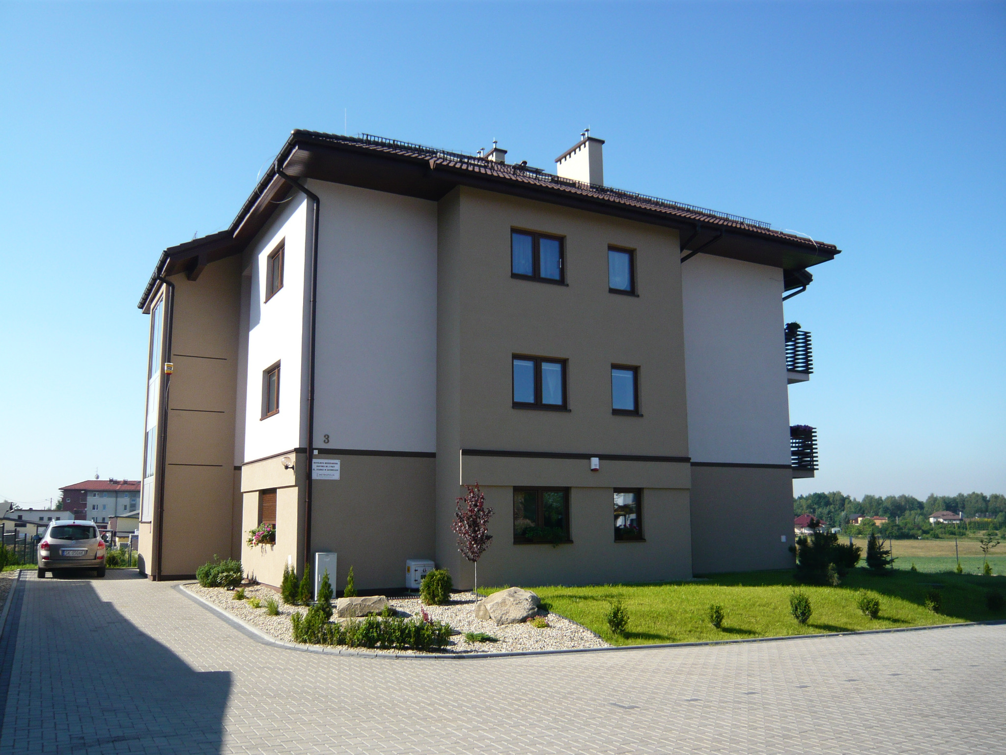 Trzypiętrowy budynek mieszkalny z beżowo-białą elewacją, brązowym dachem i oknami, otoczony zielenią, z samochodem zaparkowanym obok.
