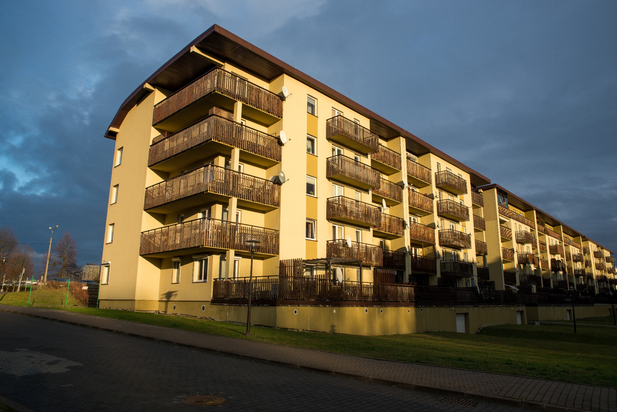 Żółty blok mieszkalny z drewnianymi balkonami pod pochmurnym niebem, widok z poziomu chodnika.