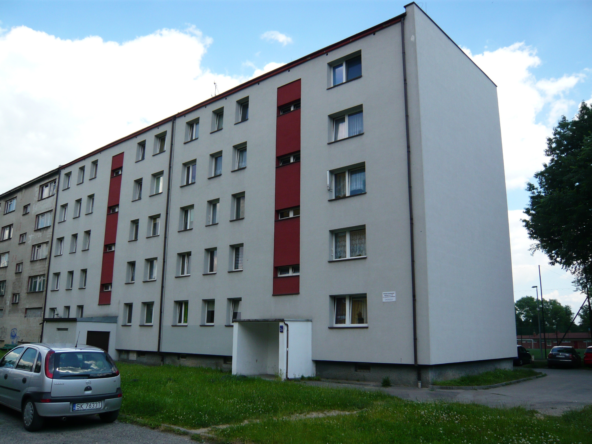 Wielopiętrowy blok mieszkalny z szarą elewacją, czerwonymi akcentami i widocznym numerem budynku '3A' nad wejściem, z zaparkowanym srebrnym samochodem osobowym na pierwszym planie.