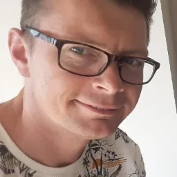 Portret uśmiechniętego brokera ubezpieczeniowego z Gniezna w okularach, w koszulce z motywem tropikalnym, na jasnym tle.