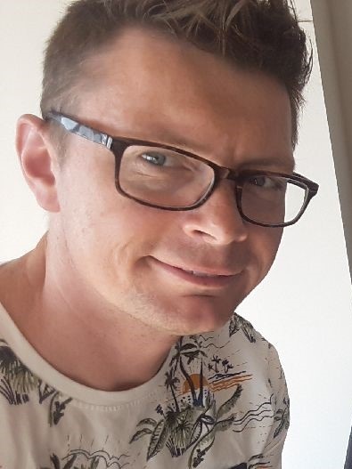 Portret uśmiechniętego brokera ubezpieczeniowego z Gniezna w okularach, w koszulce z motywem tropikalnym, na jasnym tle.