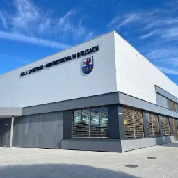 Nowoczesna hala sportowo-widowiskowa z jasną elewacją panelową i szarym obramowaniem okien, na której widnieje herb miasta Brusy na tle błękitnego nieba z delikatnymi obłokami.