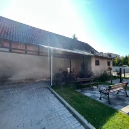 Stary budynek z szarą, falistą płytą na dachu, widoczne fragmenty muru pruskiego, obok zaparkowany samochód i elementy siłowni zewnętrznej.