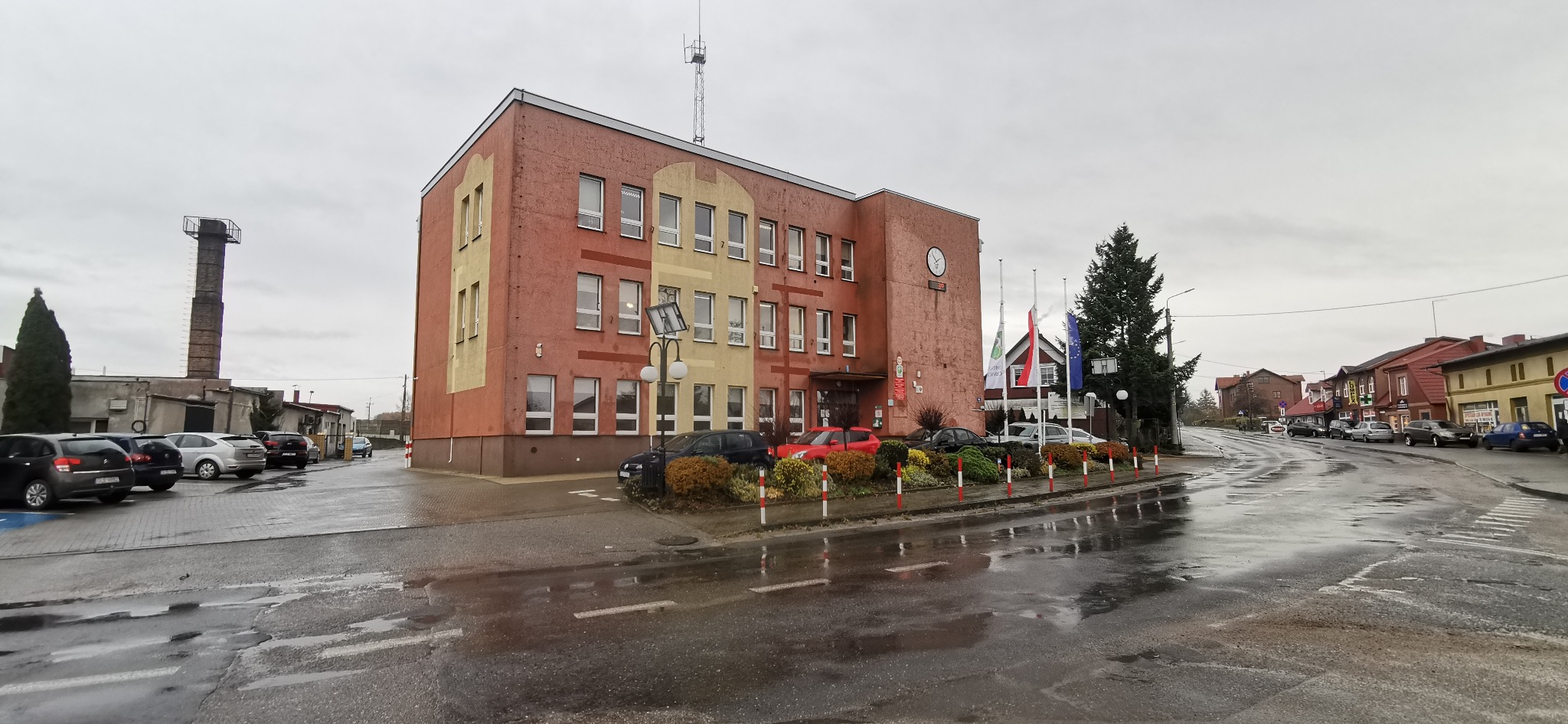 Dwupiętrowy budynek administracyjny z cegły w odcieniach czerwieni i beżu, z zegarem na fasadzie, otoczony zadbaną zielenią i flagami, widoczny z mokrej, odbijającej światło ulicy w pochmurny dzień.