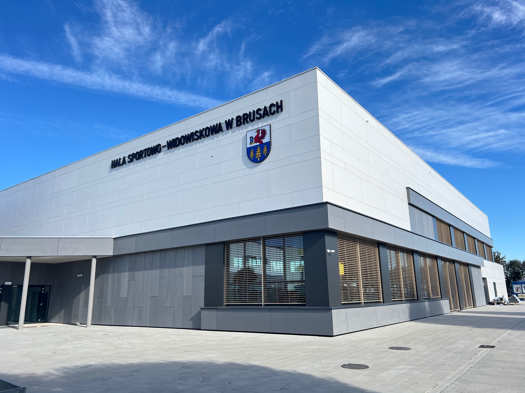 Nowoczesna hala sportowo-widowiskowa z jasną elewacją panelową i szarym obramowaniem okien, na której widnieje herb miasta Brusy na tle błękitnego nieba z delikatnymi obłokami.