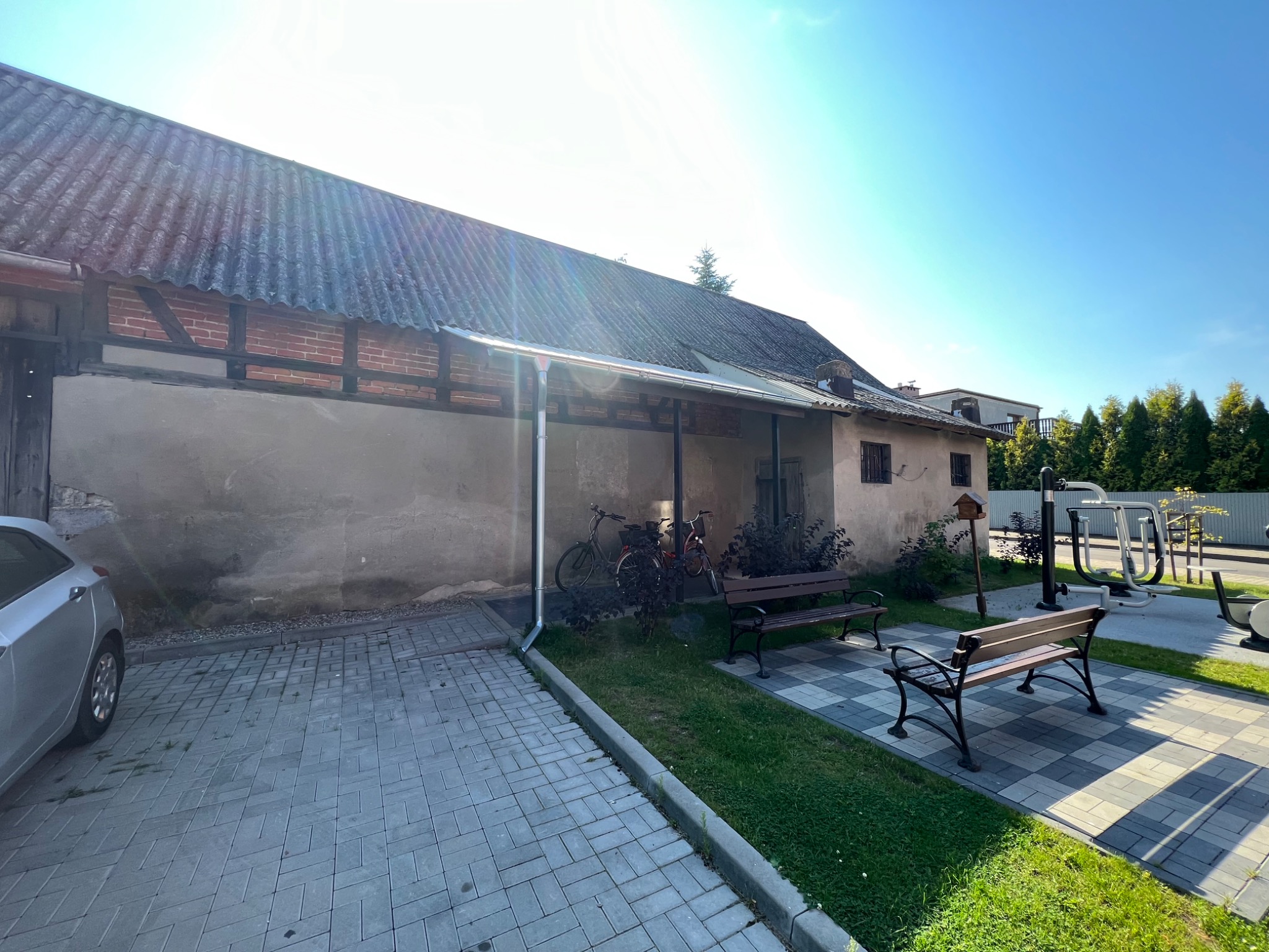 Stary budynek z szarą, falistą płytą na dachu, widoczne fragmenty muru pruskiego, obok zaparkowany samochód i elementy siłowni zewnętrznej.