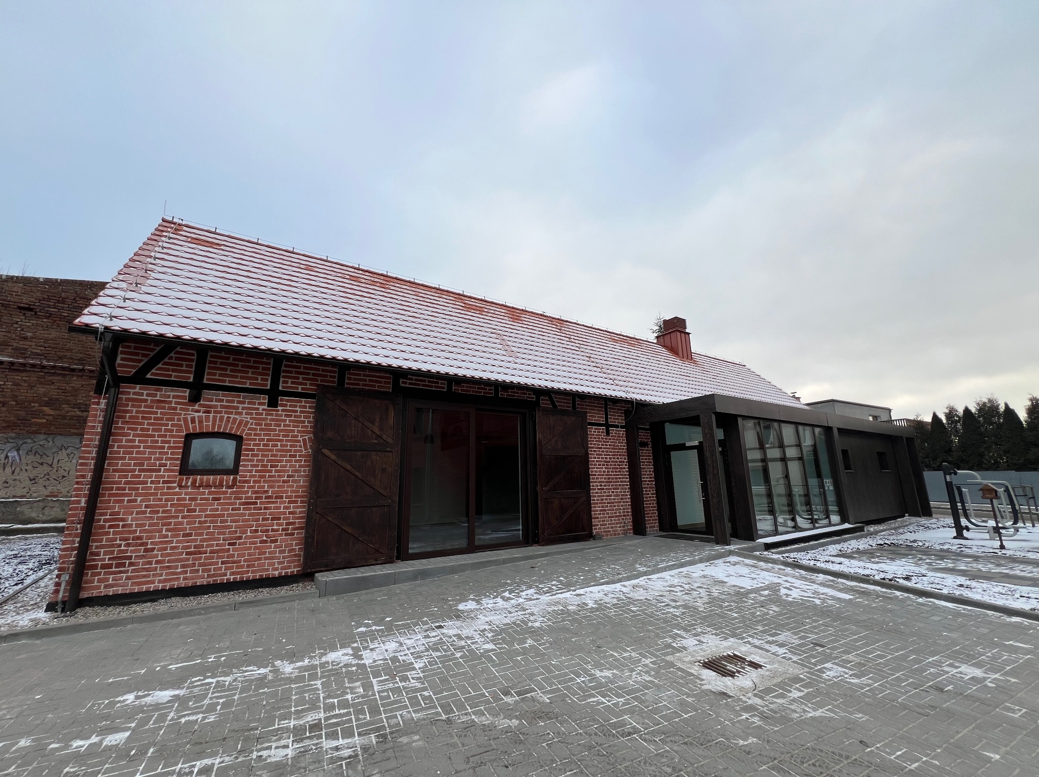 Nowoczesny budynek z czerwonej cegły z elementami drewnianymi, częściowo pokryty śniegiem, z dużymi drewnianymi wrotami i przeszkloną dobudówką, widoczny z zewnątrz na tle pochmurnego nieba.