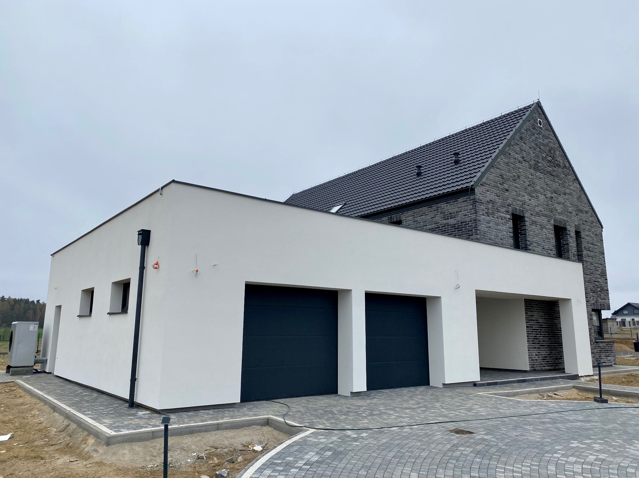 Nowoczesny dom jednorodzinny z płaskim dachem nad garażem, wykończony białą elewacją i grafitową cegłą, z dwoma bramami garażowymi i brukowanym podjazdem.