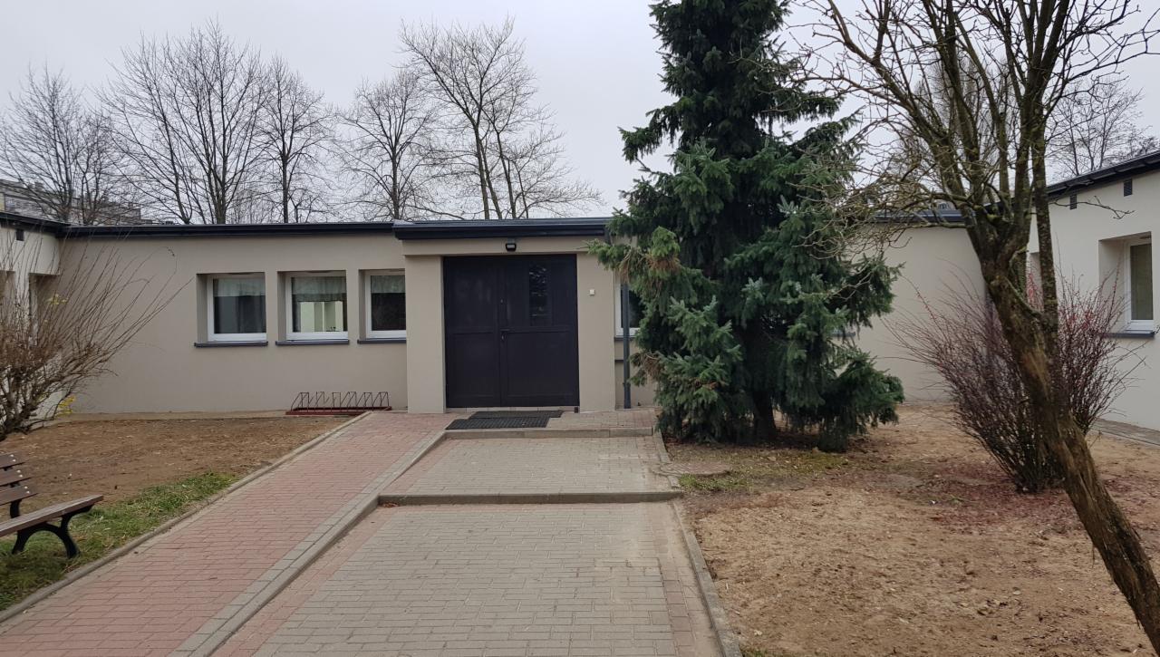 Niski, parterowy budynek z płaskim dachem, beżową elewacją i ciemnymi drzwiami wejściowymi, otoczony zielenią i wyłożony kostką brukową. Widoczne okna z białymi ramami i stojak na rowery.