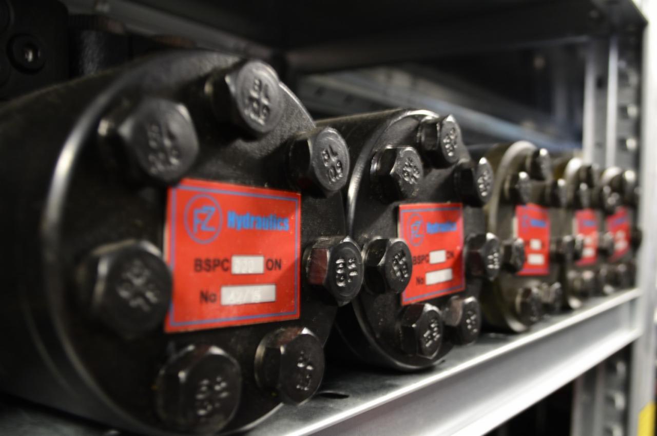 Czarne pompy hydrauliczne z czerwonymi etykietami, ustawione rzędem na metalowej półce w magazynie.