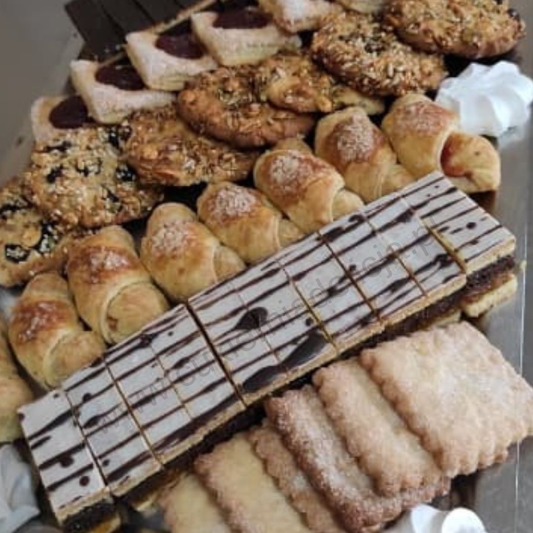 Różnorodny asortyment wypieków: rogaliki z nadzieniem, ciastka z dżemem, kwadratowe ciastka z polewą i ciastka posypane orzechami, ułożone na metalowej tacy.