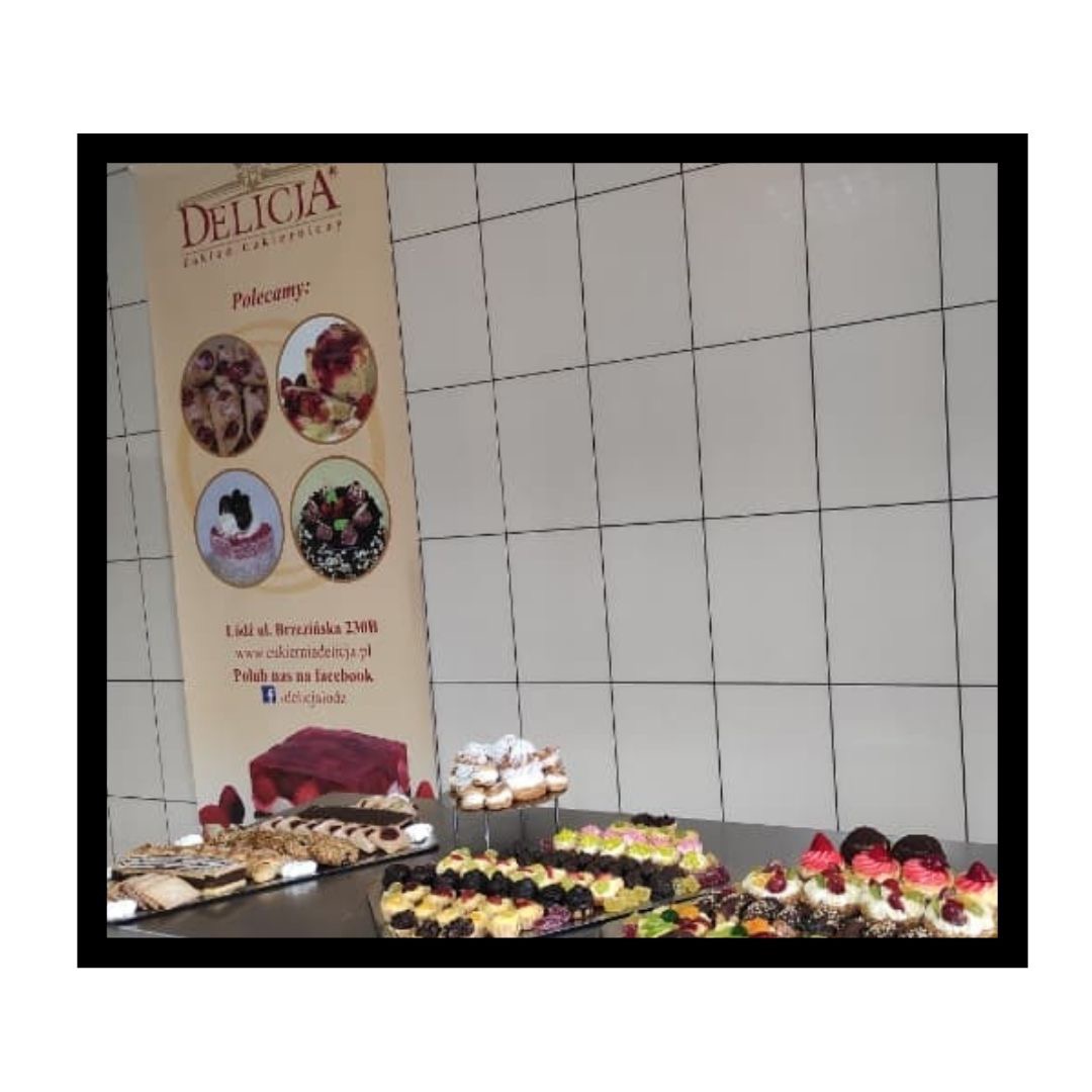 Różnorodny wybór ciast i ciasteczek na ladzie cukierniczej, z plakatem reklamowym 'Delicia' w tle.