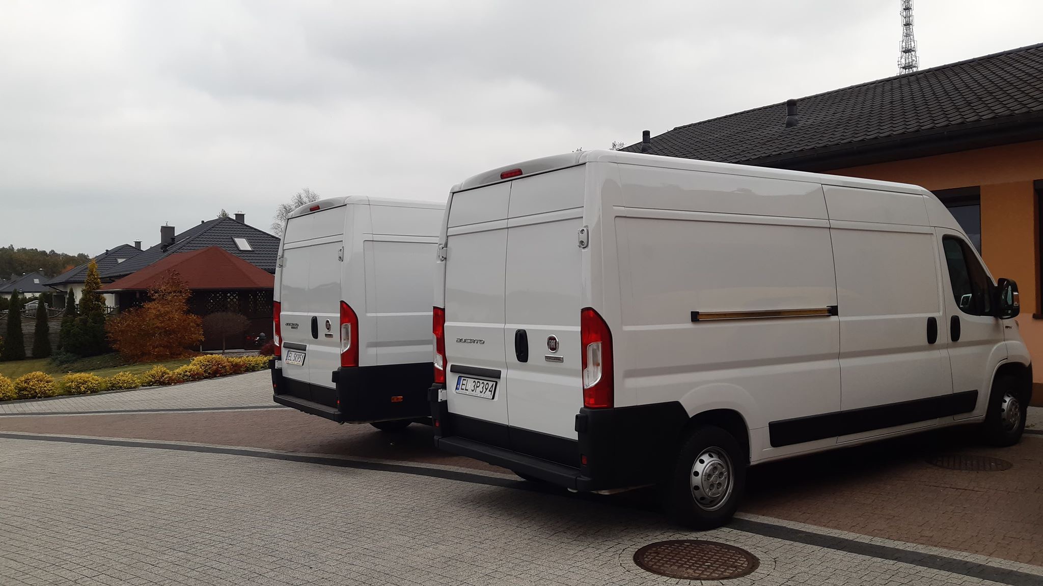 Dwa białe busy Fiat Ducato zaparkowane na kostce brukowej przed domem z brązowym dachem i pomarańczową elewacją w pochmurny dzień.