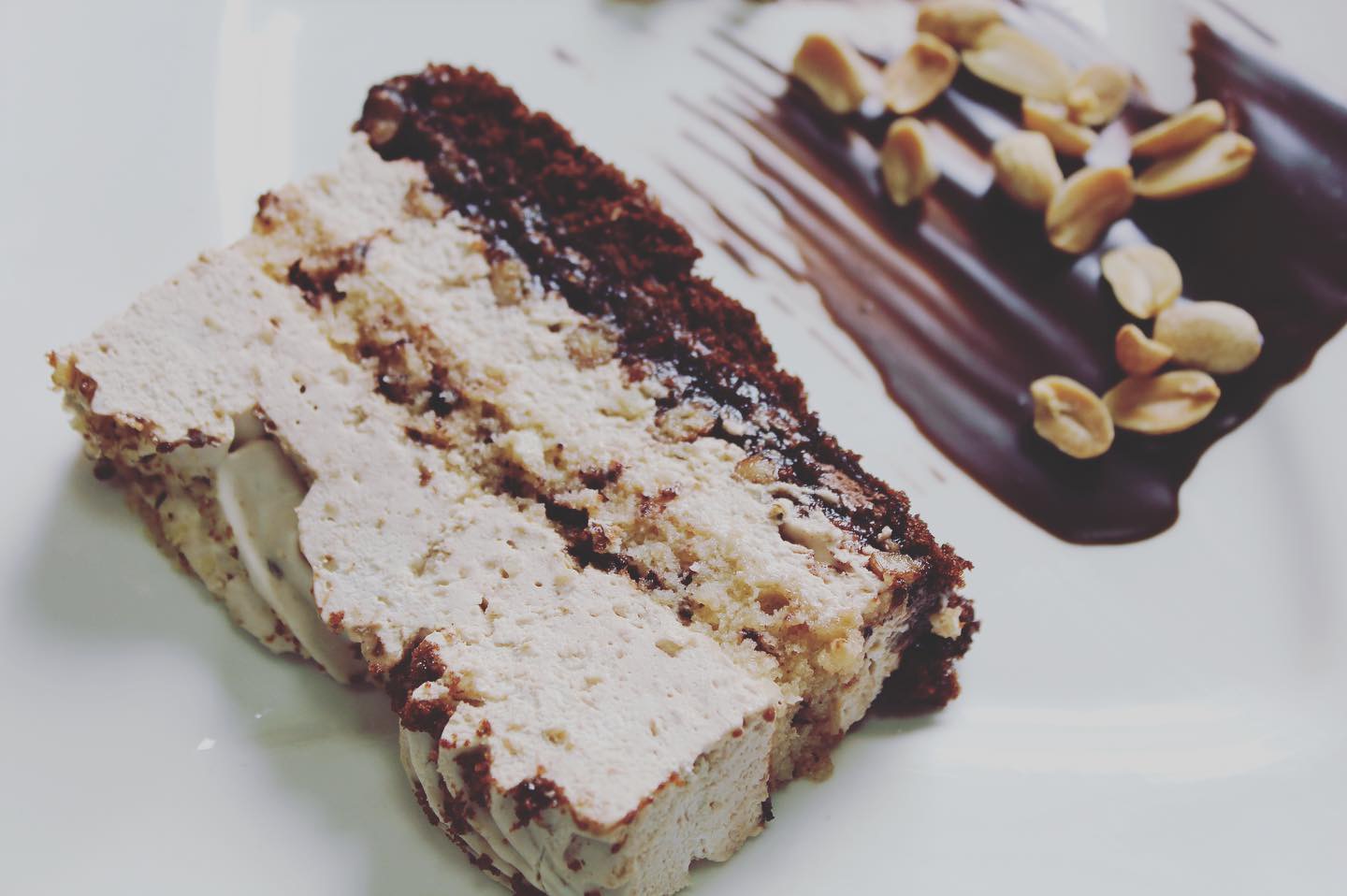 Kawałek tortu z kremem kawowym, ciemnym biszkoptem i polewą czekoladową z orzechami arachidowymi na białym talerzu.