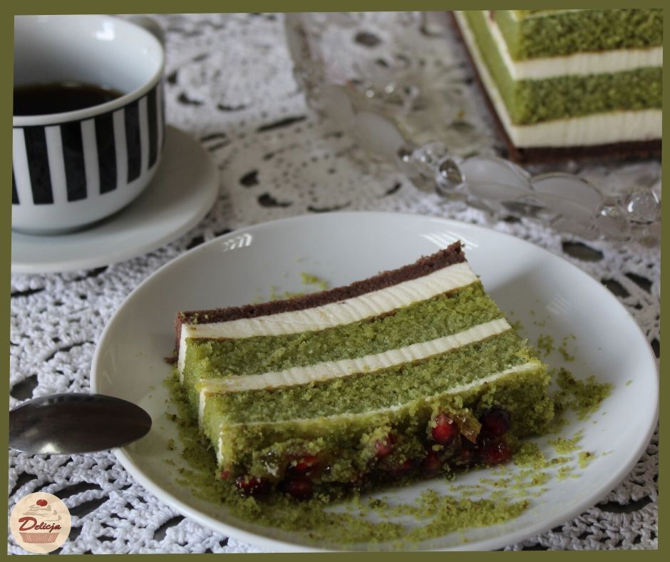 Kawałek zielonego tortu warstwowego z kremem, posypany kruszonką i owocami granatu na białym talerzu, obok filiżanka kawy w czarno-białe paski na koronkowej serwetce.
