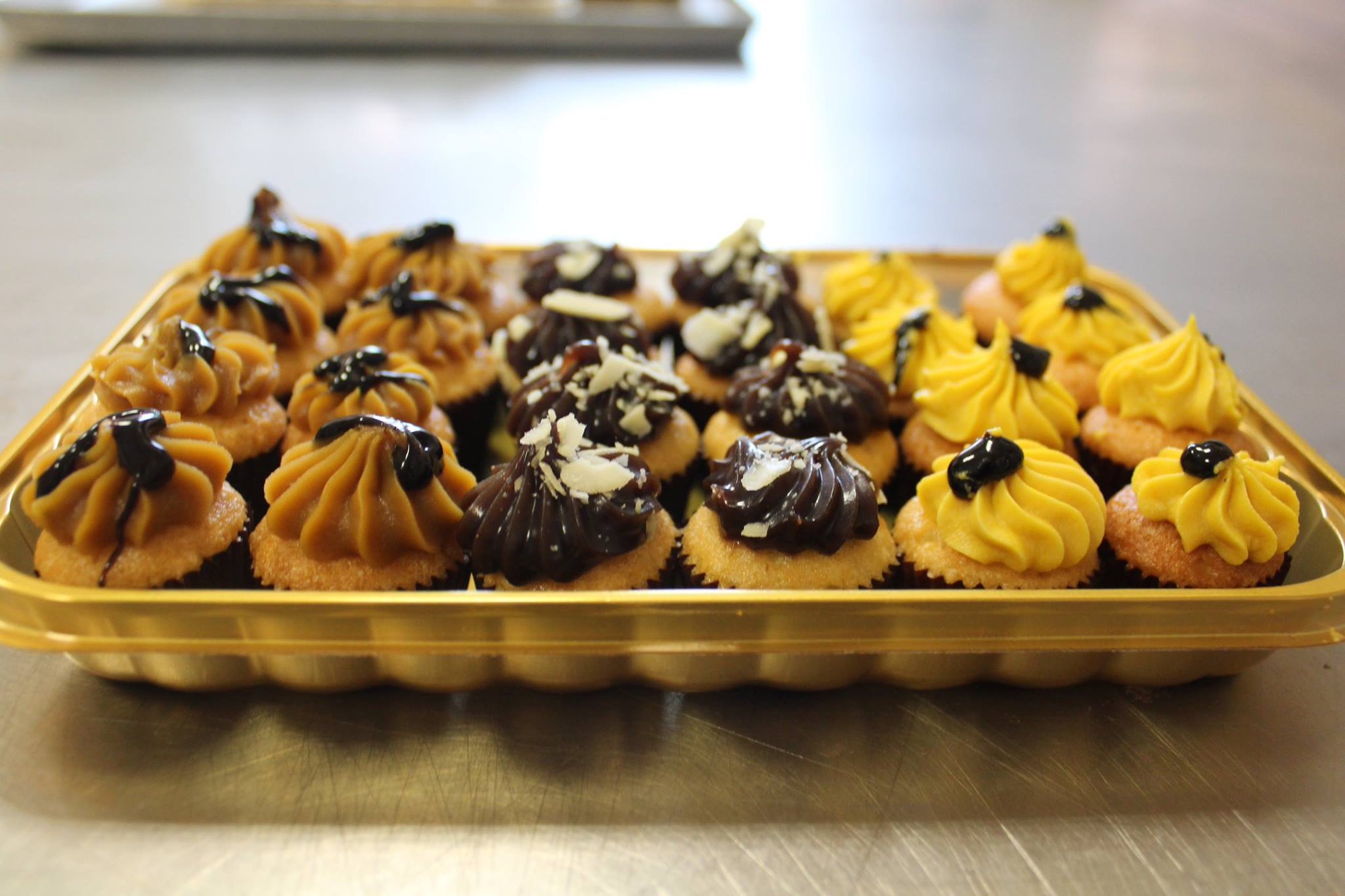 Rząd babeczek na złotej tacy: krem karmelowy z polewą, czekoladowy z wiórkami kokosowymi i żółty z czarną kropką.
