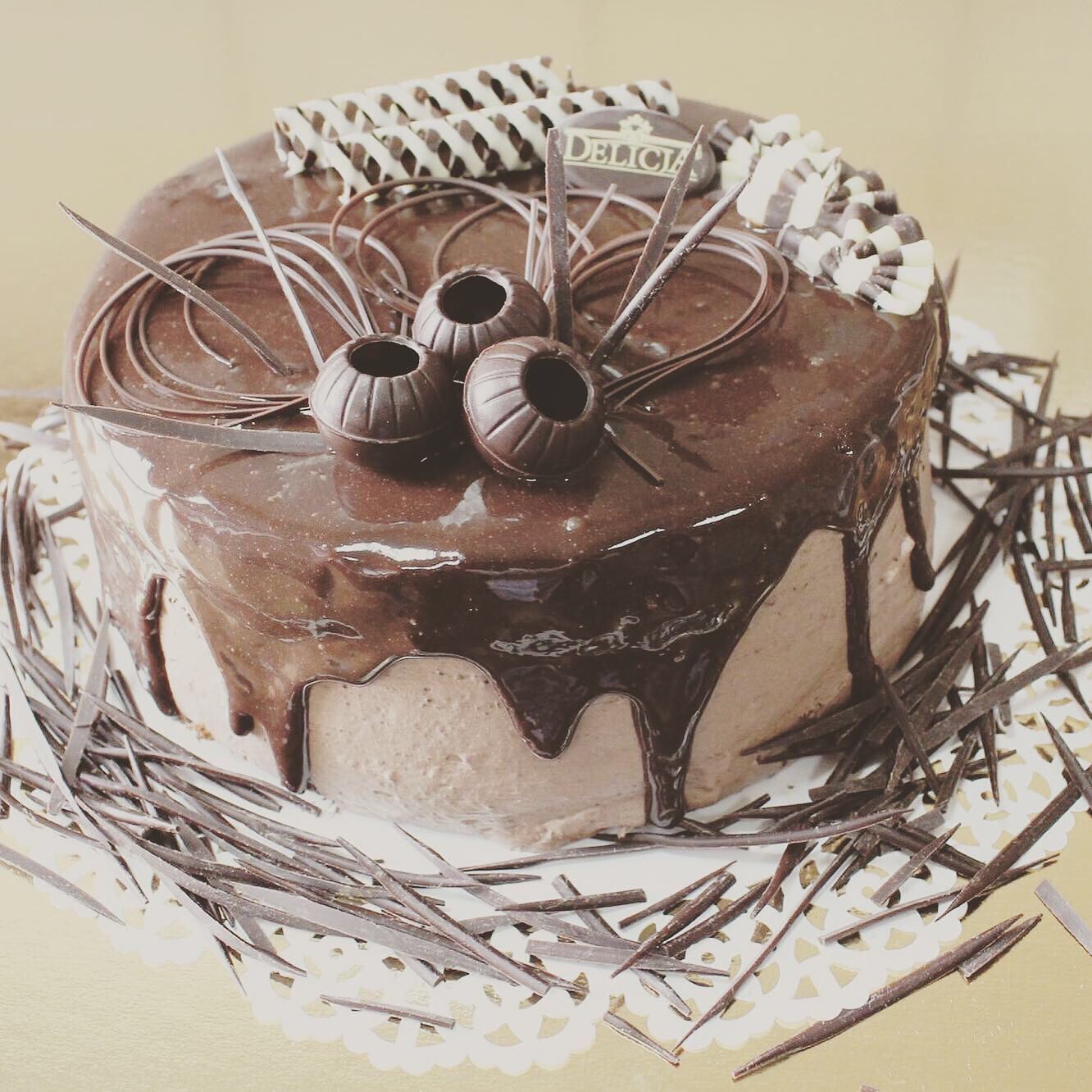 Czekoladowy tort z polewą, ozdobiony kulkami czekoladowymi z wgłębieniami, czekoladowymi wiórkami i dekoracją z białej i ciemnej czekolady, umieszczony na koronkowej podkładce.