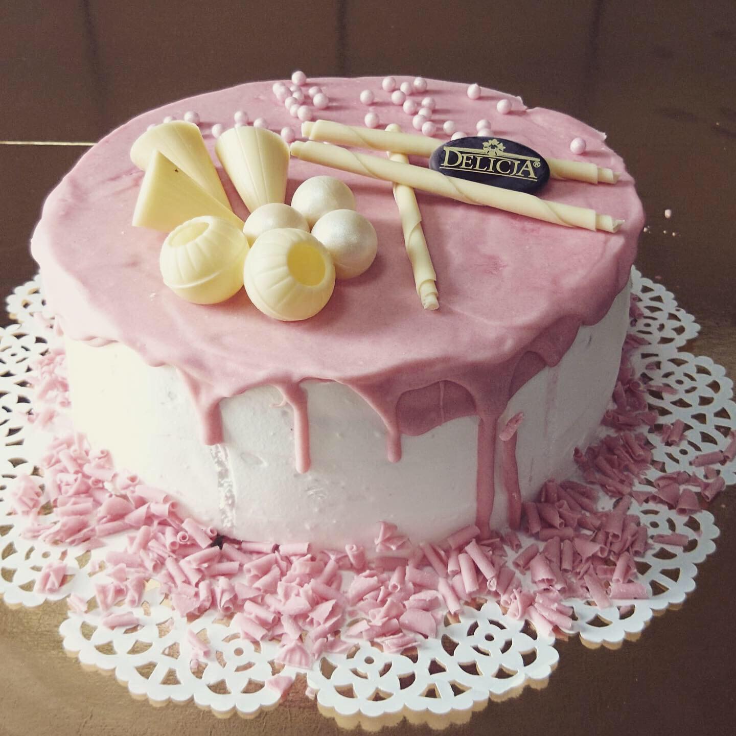 Okrągły tort z białym kremem i różową polewą, ozdobiony białymi czekoladowymi kulkami, rurkami, stożkami i posypką, z logo 'Delicia' na wierzchu, umieszczony na białej koronkowej podkładce.