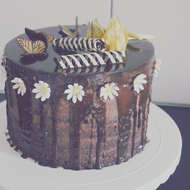 Czekoladowy tort z polewą, dekorowany białymi kwiatkami, czekoladowymi rulonikami i motylkiem, wykonany w Łodzi.