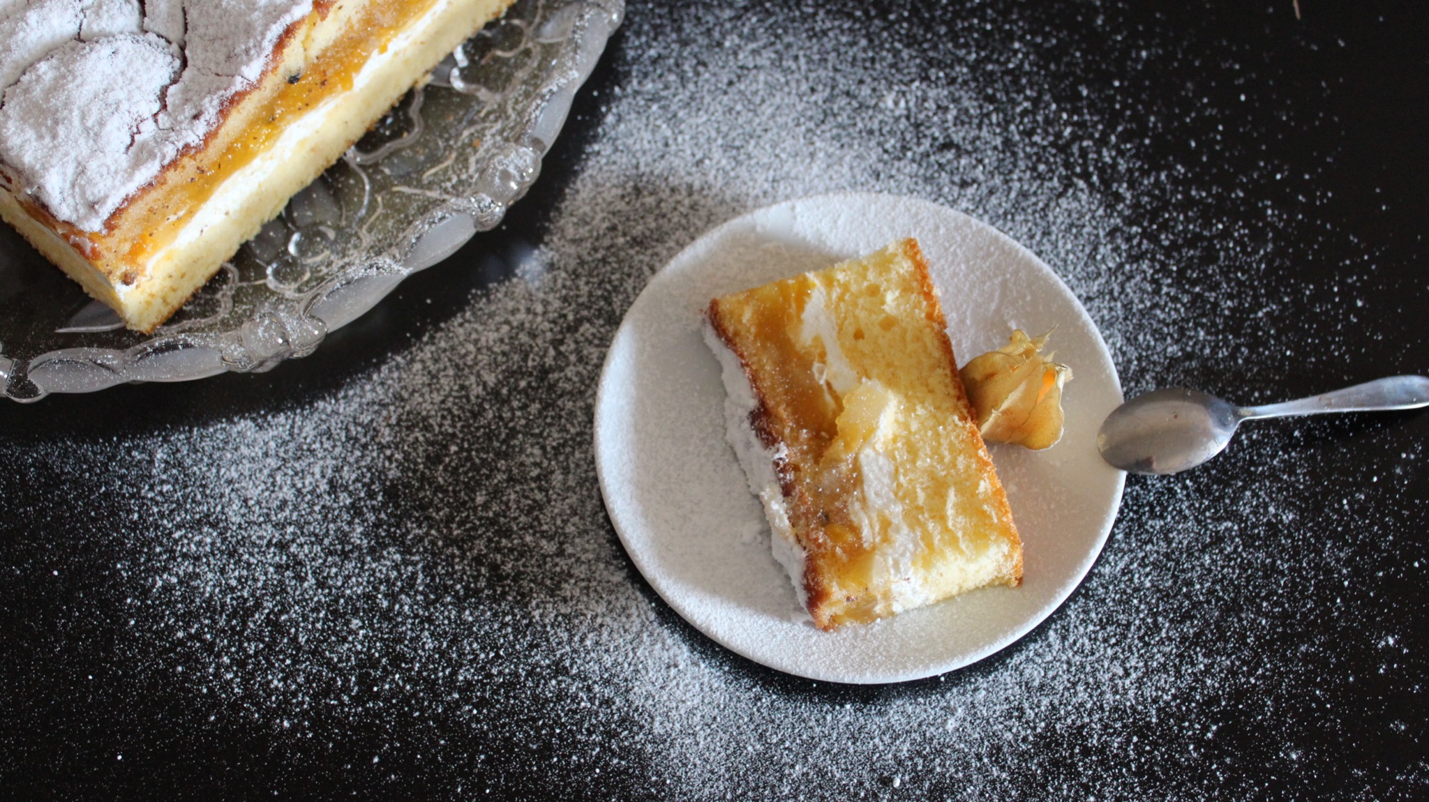 Kawałek ciasta z kremem i galaretką, posypany cukrem pudrem, na białym talerzyku z łyżeczką i miechunką peruwiańską, obok większy kawałek na szklanym talerzu, całość na czarnym tle posypanym cukrem...