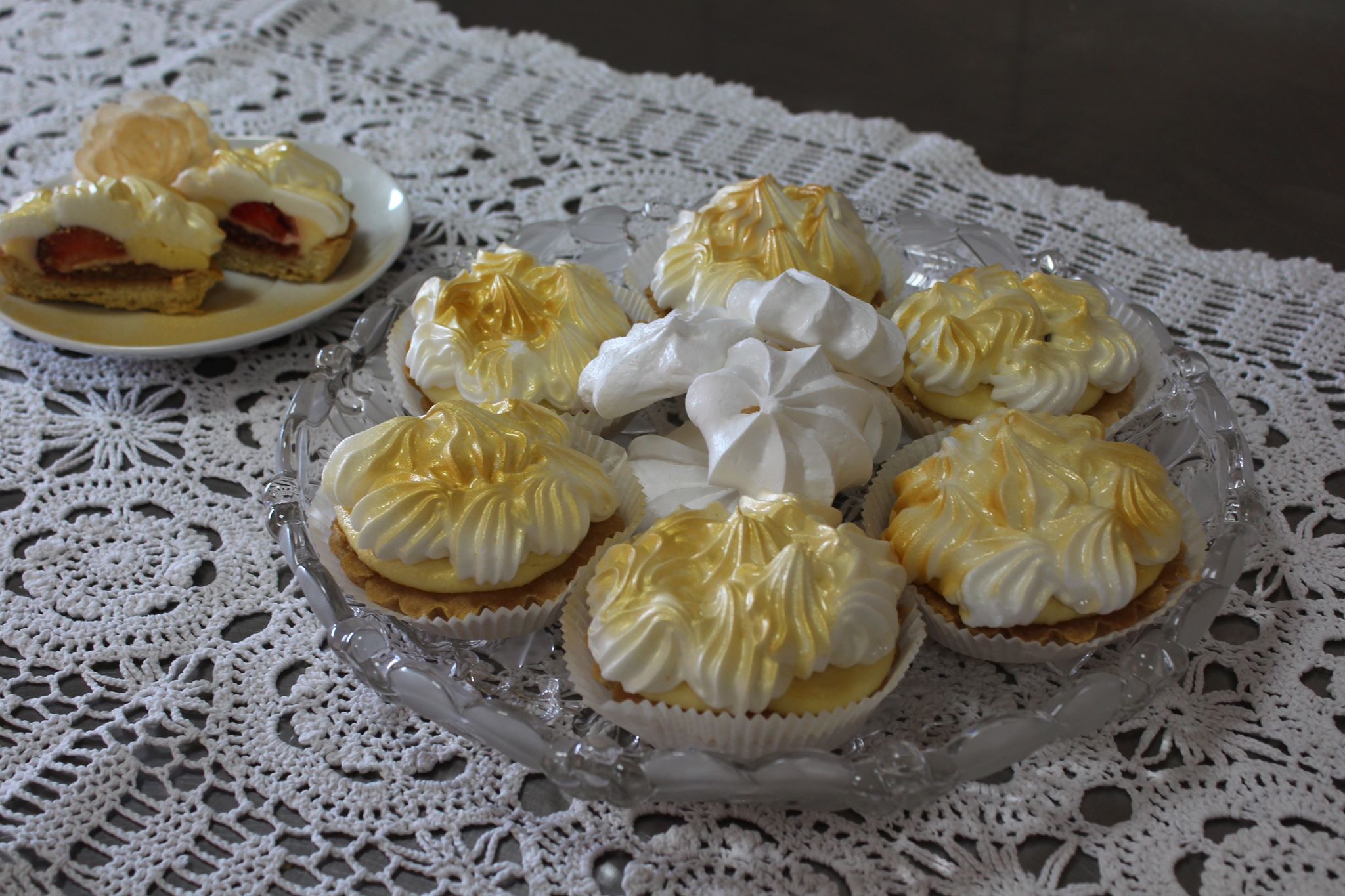 Talerz z deserami: tartaletki z kremem cytrynowym i bezą muśniętą złotem, obok tartaletki z truskawkami, na białej, ażurowej serwecie.