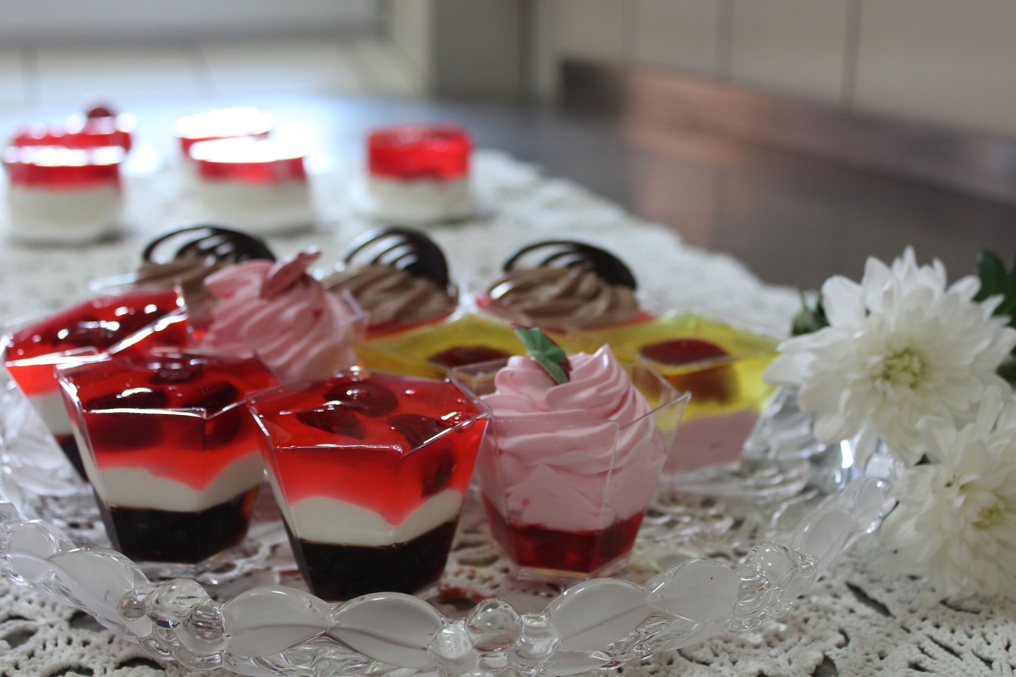 Deserowe pucharki z galaretką, kremem i owocami na ażurowej tacy, dekorowane białymi kwiatami.