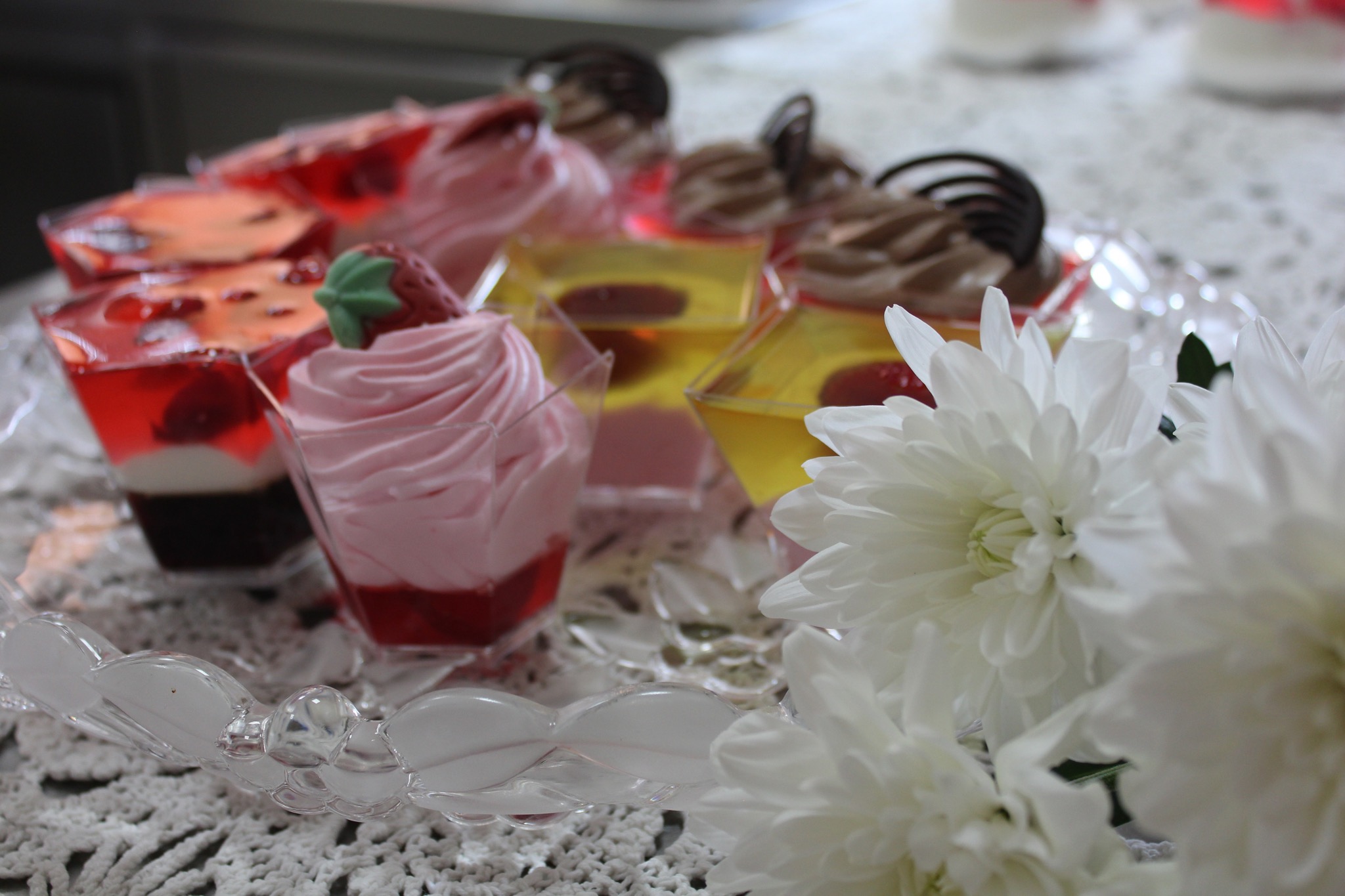 Deserki w przezroczystych naczyniach na szklanej tacy, z dekoracją z białych kwiatów, na białym obrusie z koronki.