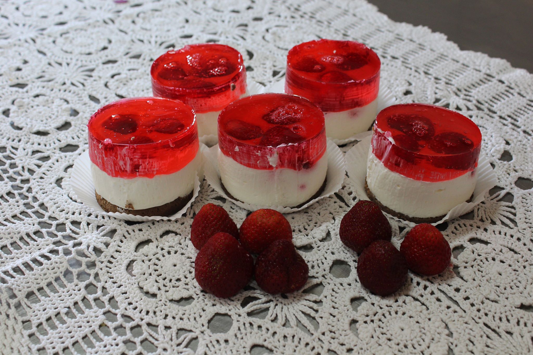 Pięć mini serników z galaretką truskawkową i świeżymi truskawkami na białej, ażurowej serwetce.