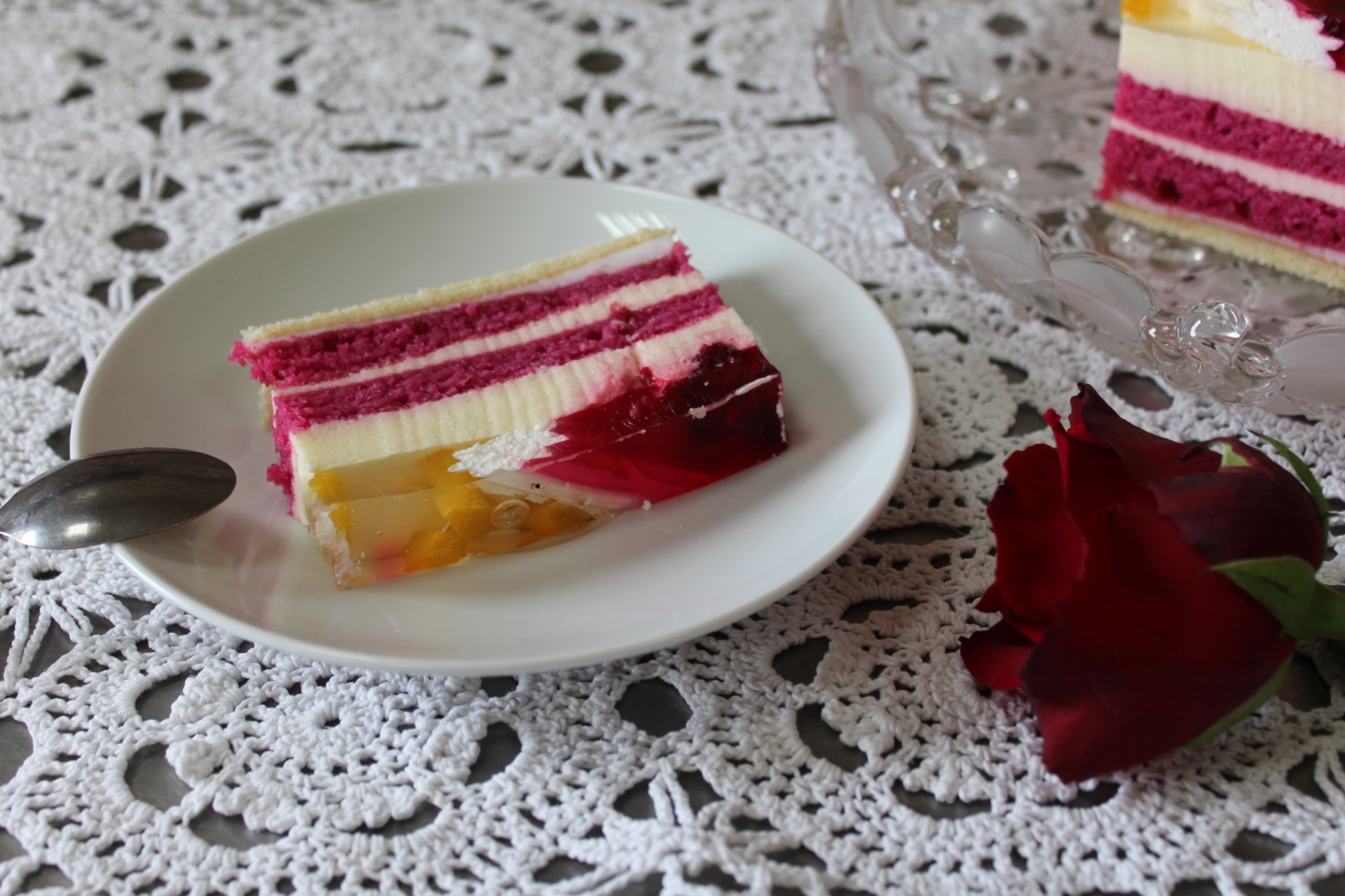 Kawałek warstwowego ciasta z różowymi biszkoptami, kremem i galaretką owocową na białym talerzu obok srebrnej łyżeczki, na białym, ażurowym obrusie, z czerwoną różą w tle.
