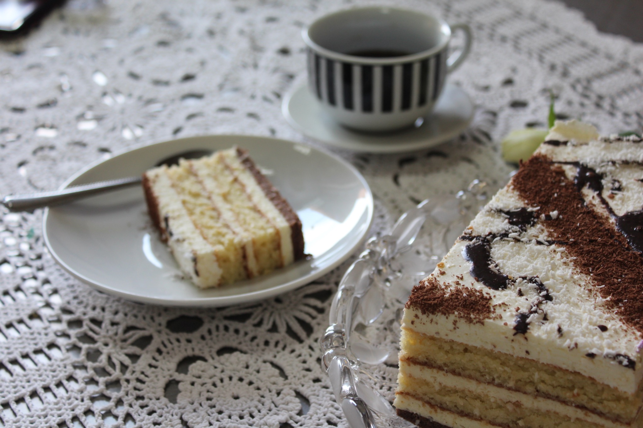 Kawa w czarno-białym kubku i dwa kawałki tortu na talerzach, dekorowane czekoladą i wiórkami kokosowymi, na białym, koronkowym obrusie.