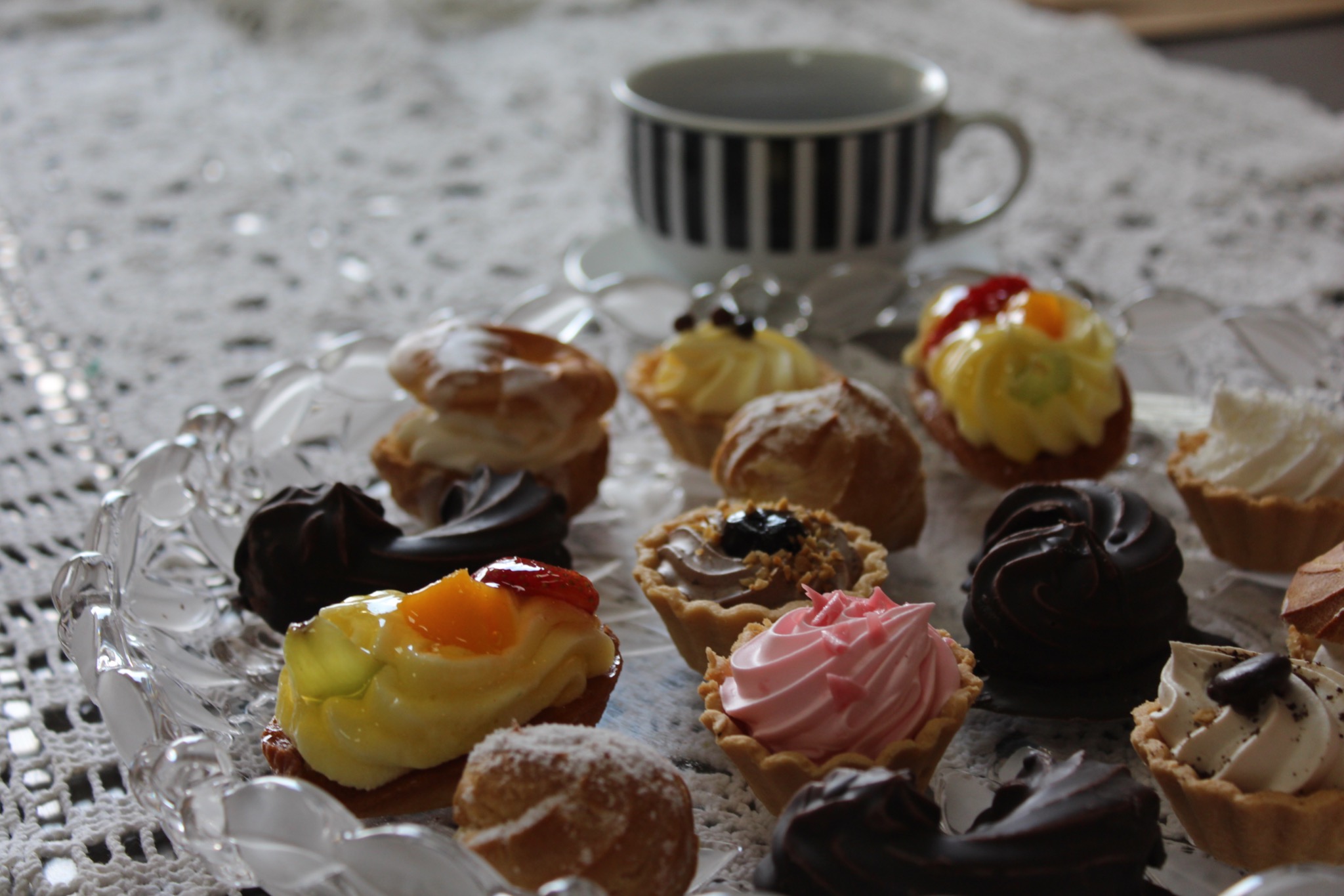 Szklany talerz z różnorodnymi mini ciasteczkami i babeczkami, w tle filiżanka w czarno-białe paski na białym, ażurowym obrusie.
