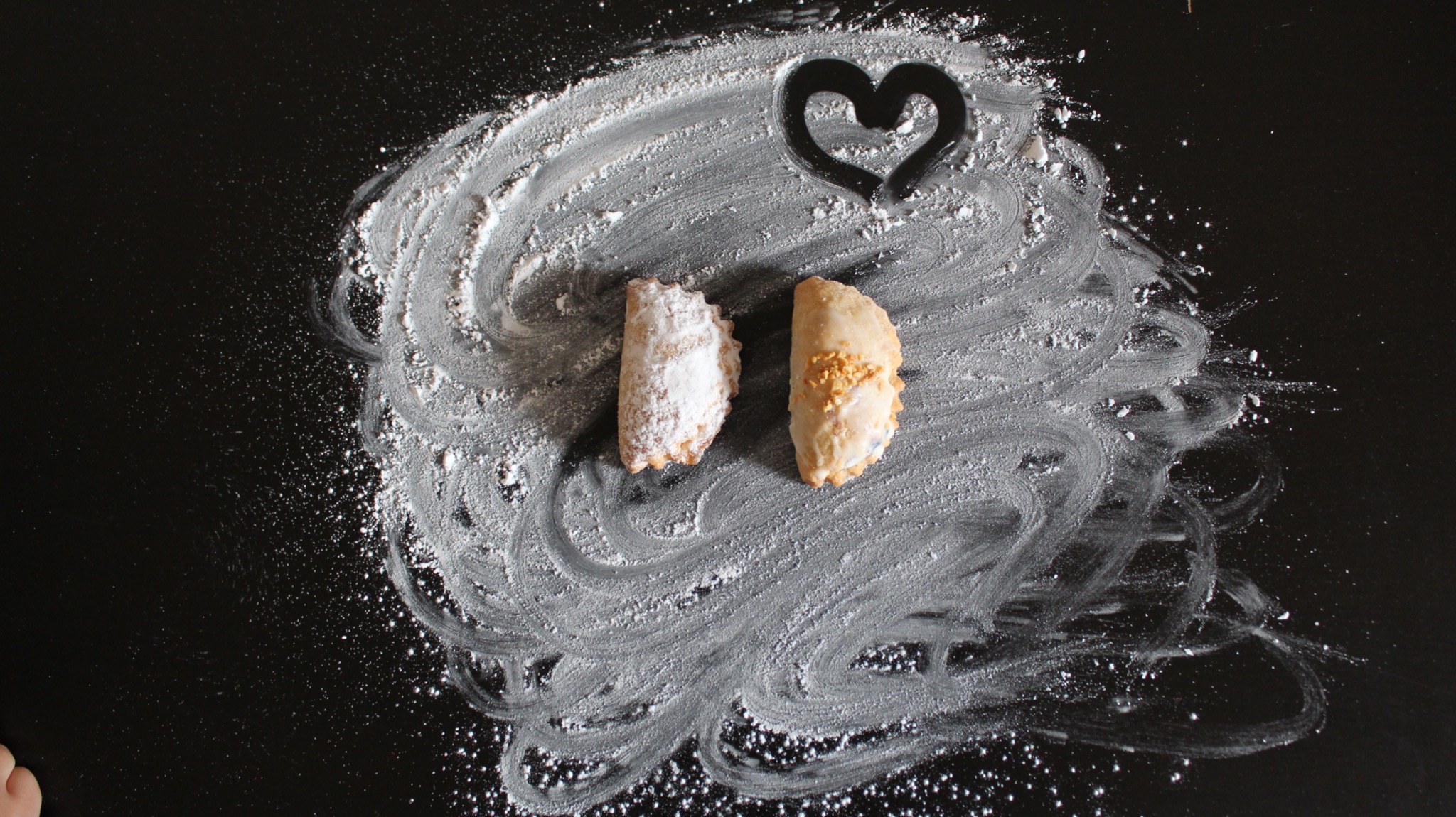 Dwa pierogi, jeden posypany cukrem pudrem, drugi z polewą, leżące na czarnym tle z mąką uformowaną w kształt serca.