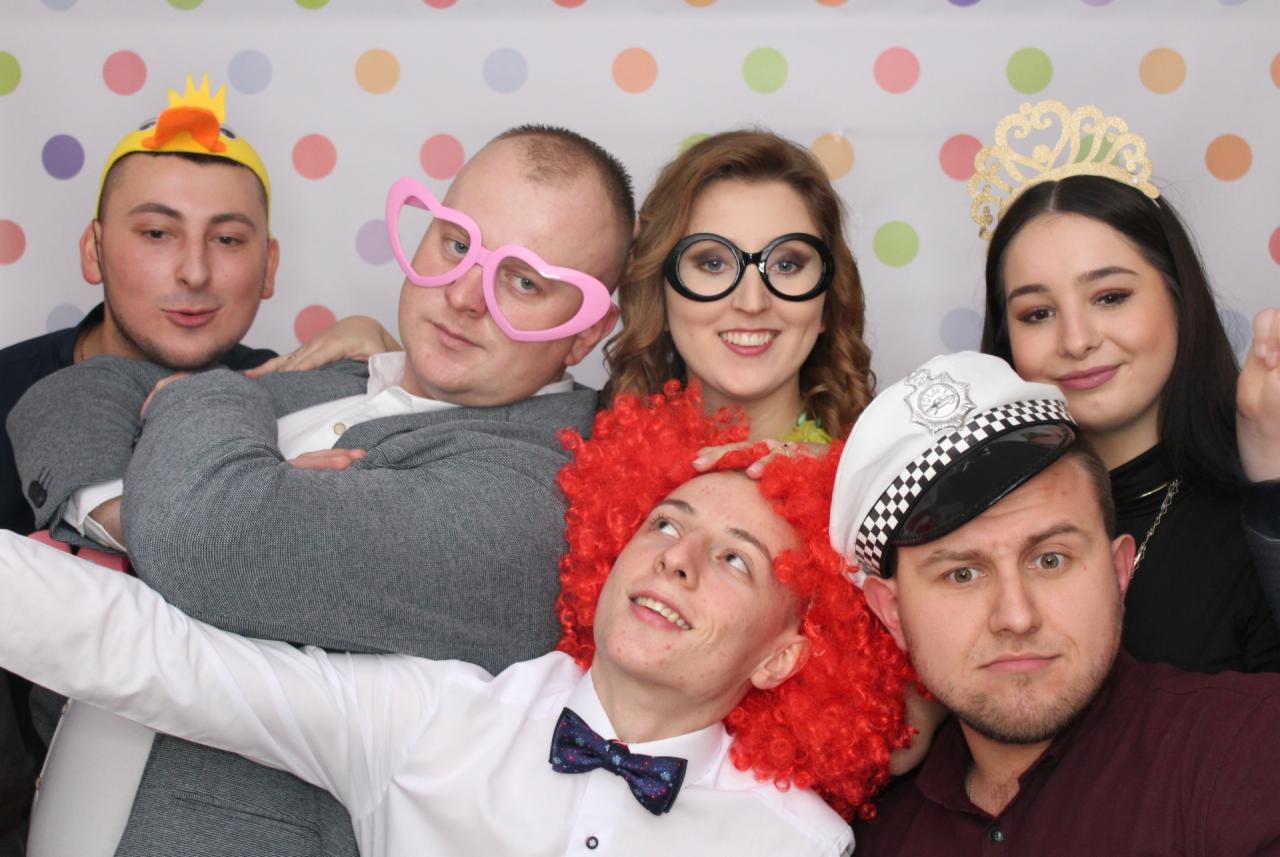 Grupa sześciu osób pozuje w przebraniach przed tłem w kolorowe kropki, w tym korony, peruka i okulary w kształcie serc.
