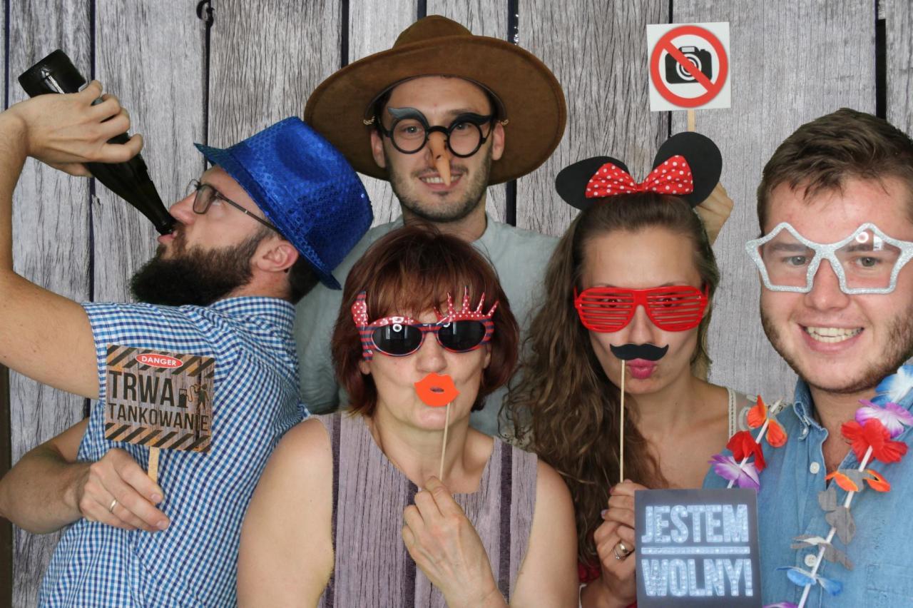 Grupa osób pozuje do zdjęcia z rekwizytami do fotobudki: śmieszne okulary, wąsy na patyku, kapelusze, tabliczki z napisami 'Trwa tankowanie' i 'Jestem wolny!' na tle drewnianej ścianki.