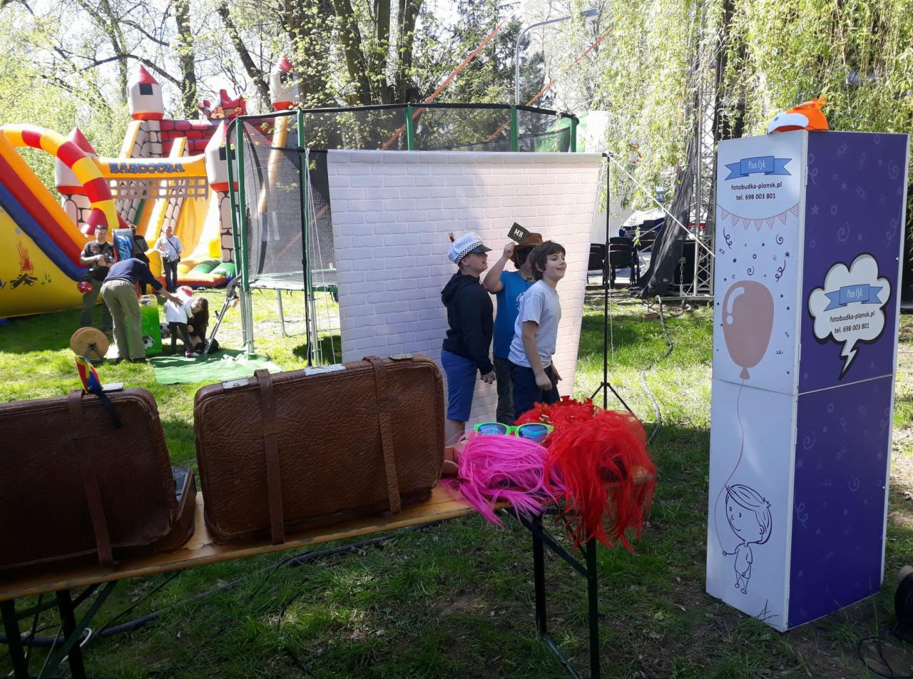 Trzech chłopców pozuje do zdjęcia na tle białej ściany z cegieł, rekwizyty: walizki, kolorowe peruki i okulary, w tle dmuchany zamek i trampolina, obok budka fotograficzna z reklamą.