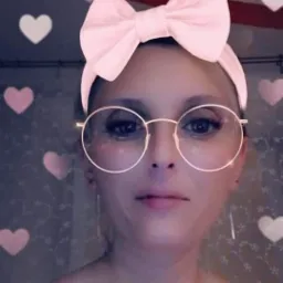 Portret kobiety w okularach z różową kokardą na głowie, na tle ściany z motywem kwiatowym i efektem serduszek.