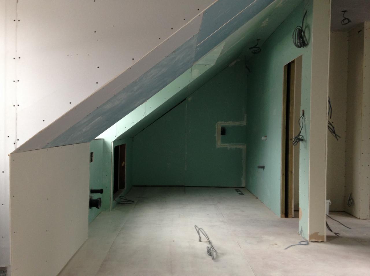 Poddasze w trakcie adaptacji: ściany wykończone płytami gipsowo-kartonowymi, widoczne otwory na instalacje elektryczne i hydrauliczne, skosy z zabudową z płyt G-K, surowy beton na podłodze.